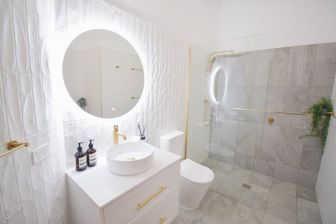 Modern white bathroom with round illuminated mirror, vessel sink, floating vanity with gold hardware, glass shower enclosure, and marble tile flooring