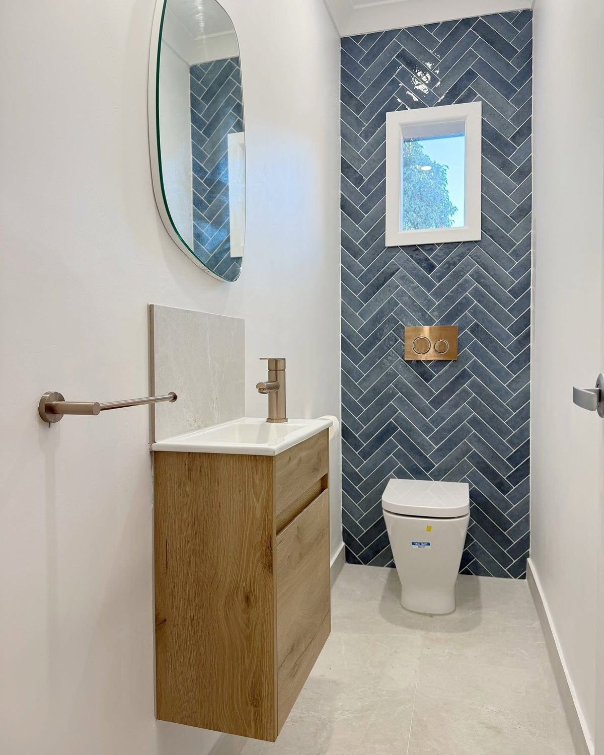 Modern bathroom with wood vanity, round mirror, dark herringbone tile accent wall, toilet, and framed artwork