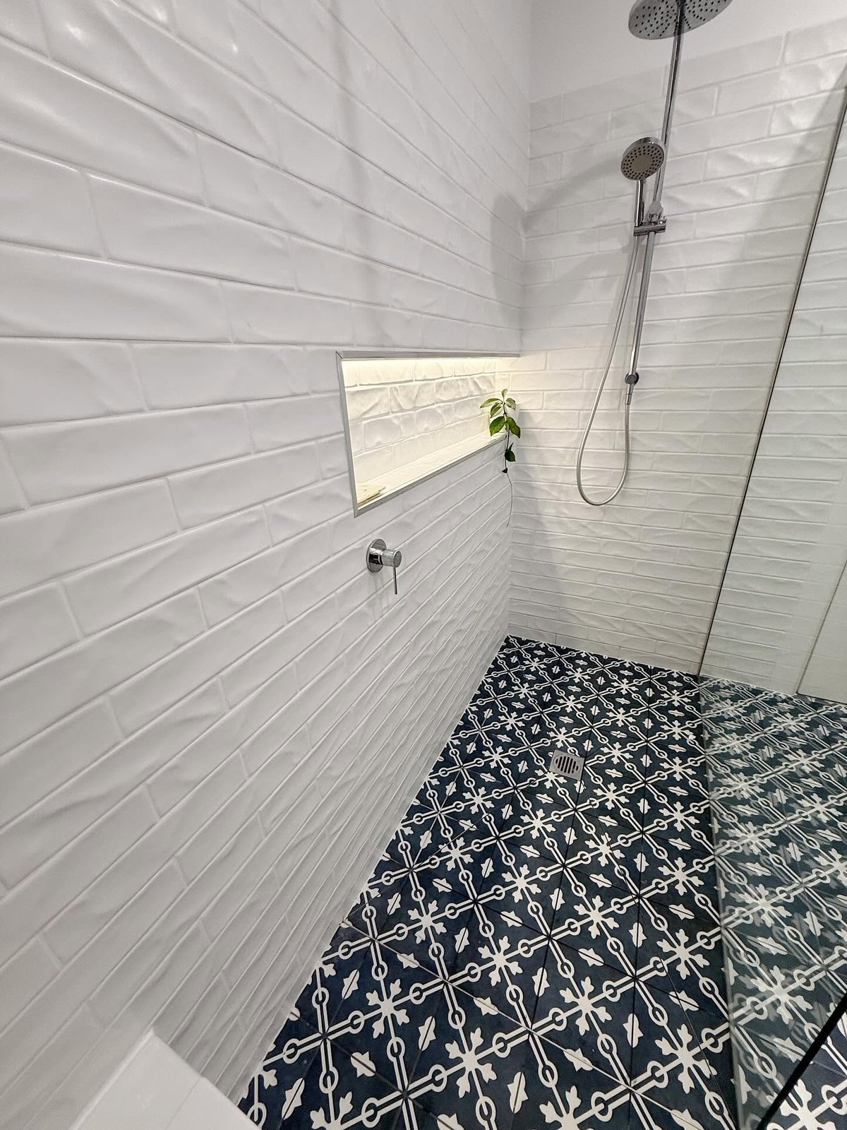 Modern shower stall with white subway tile walls, black and white patterned floor tiles, rainfall showerhead, and built-in niche with plant.