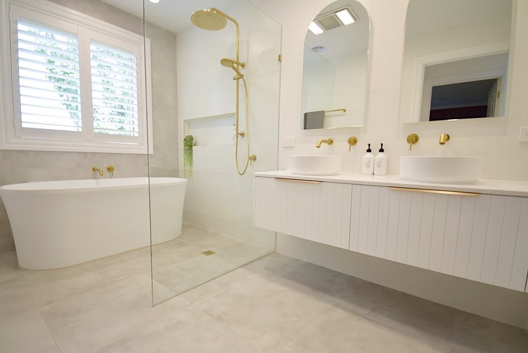 Modern bathroom with white freestanding tub, walk-in shower with glass enclosure, floating vanity with dual sinks, brass fixtures, and marble flooring.