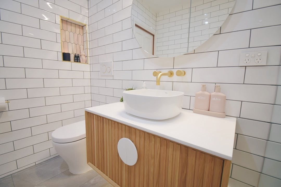 Bathroom renovation with white subway tile walls, wooden vanity with round white sink, brass faucet, toilet and pink towels