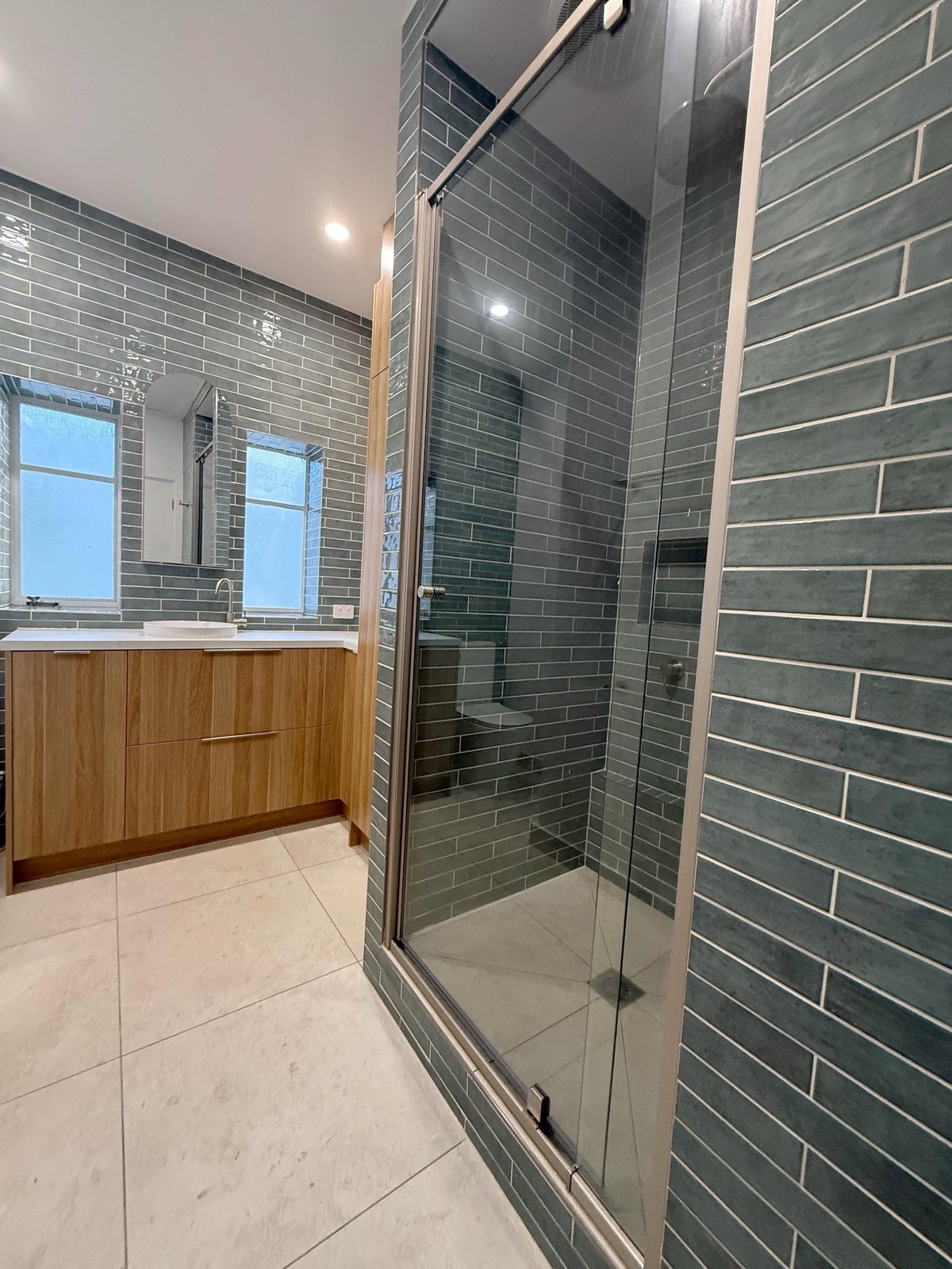 Modern bathroom with large walk-in shower, green subway tile walls, wood vanity cabinet, and light stone flooring
