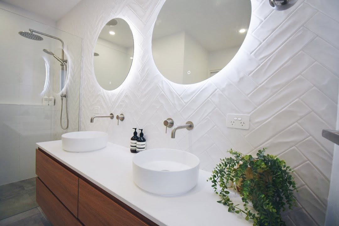 Modern white bathroom with dual round vessel sinks, illuminated mirrors, wooden vanity, white herringbone tile wall, and decorative potted plant