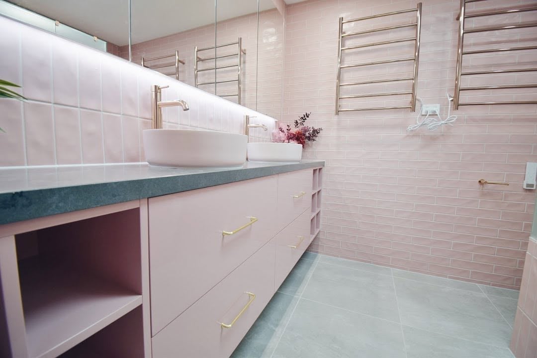 Modern bathroom with pink tile walls, white vanity, teal countertop, open shelving, and decorative louvered windows above the sink