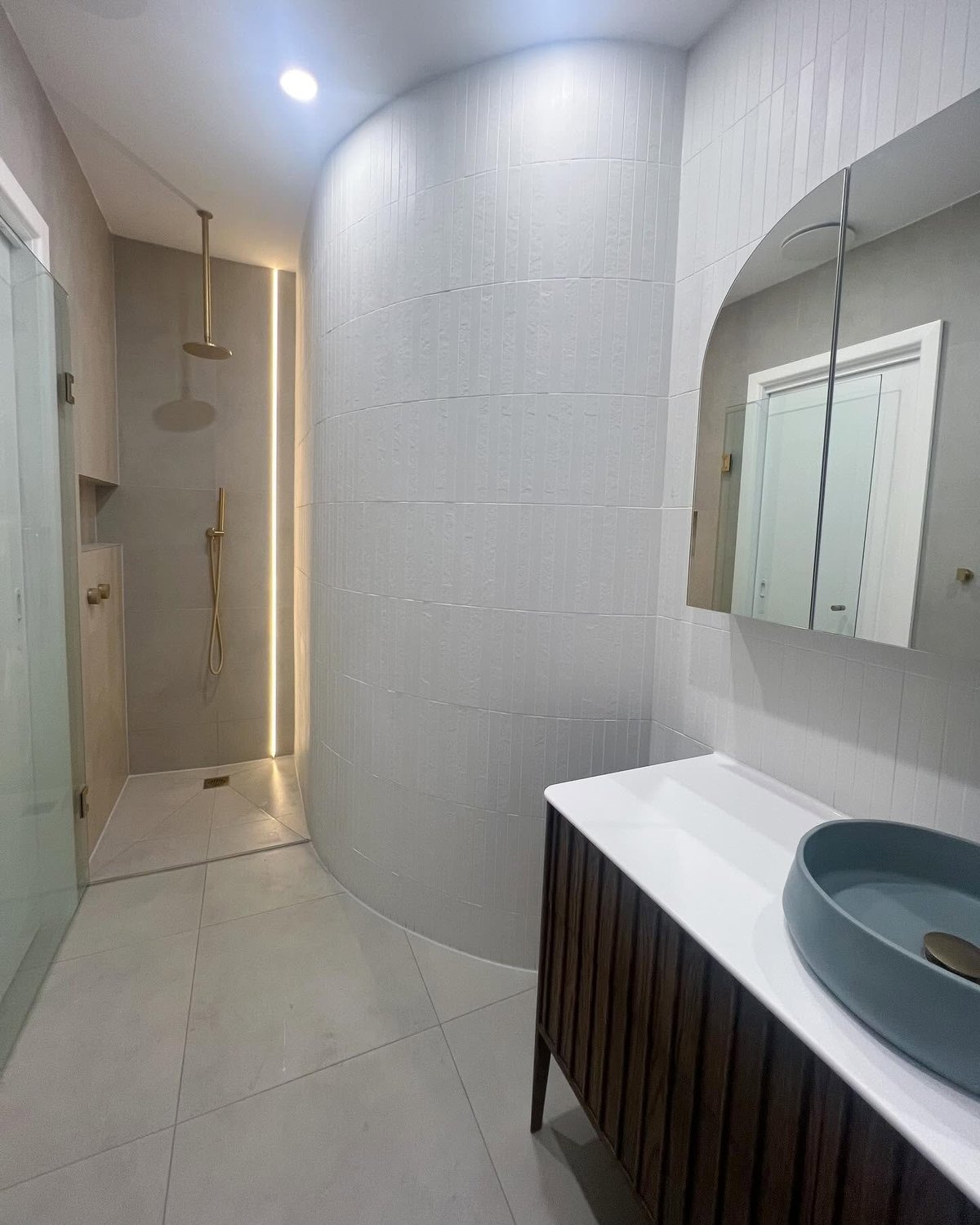 Modern bathroom interior with curved white tile walls, vanity with blue sink, illuminated mirror, and walk-in shower with glass door