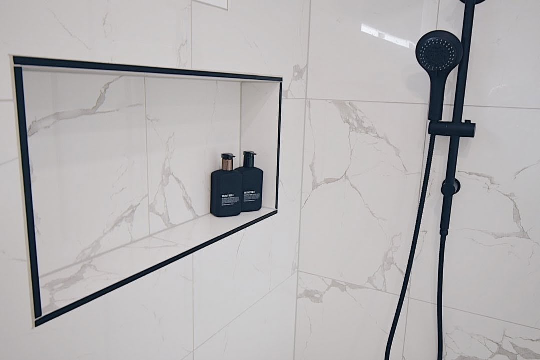 Modern shower niche with black frame holding dark bottles, next to a black rainfall showerhead on marble-tiled bathroom wall