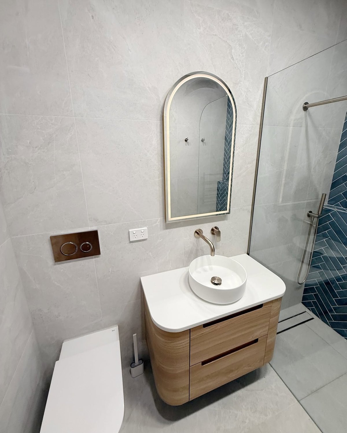 Modern bathroom vanity with white round sink, wooden cabinet, arched mirror, and glass shower enclosure with blue tile accent wall