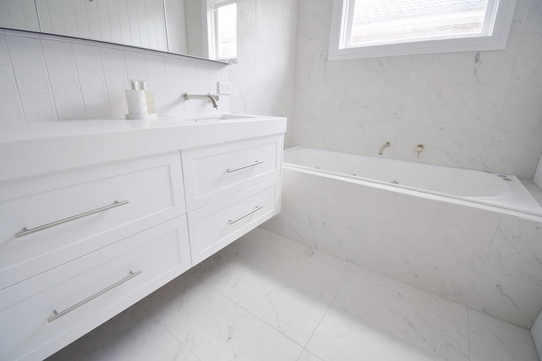 Modern white bathroom with marble tile flooring, white vanity with drawers, sink, and soaking tub near window