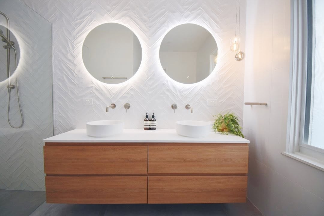 Modern bathroom vanity with two illuminated circular mirrors, white vessel sinks, wooden cabinetry, and marble tile walls