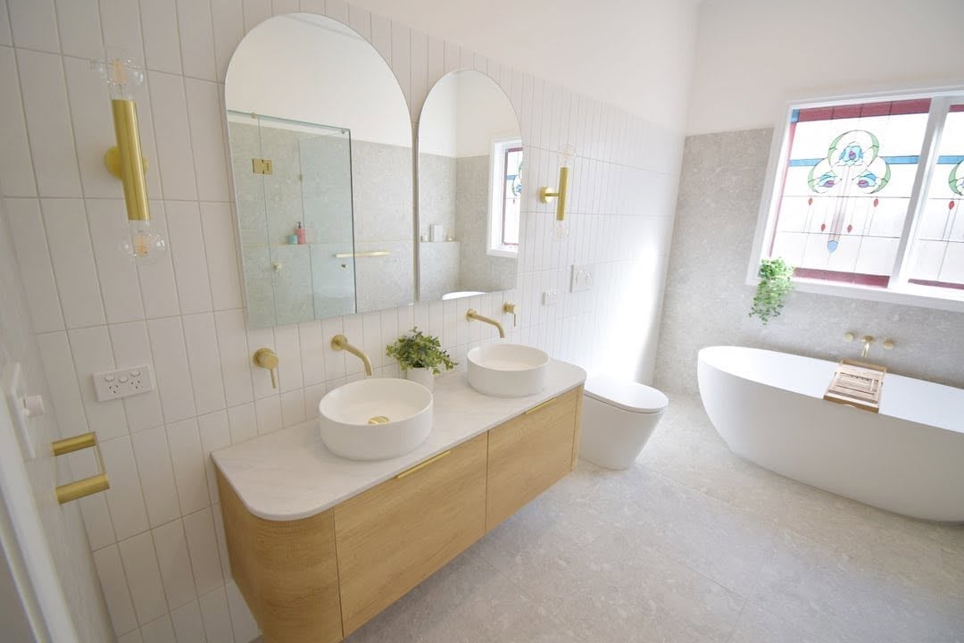 Modern bathroom with white tile walls, double vessel sinks on wooden vanity, arched mirrors, freestanding white tub, and gold fixtures