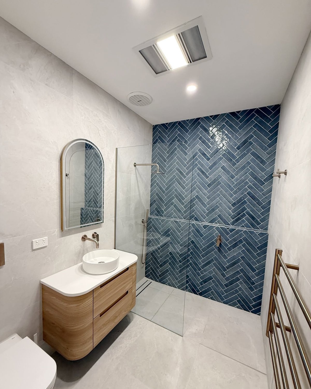 Modern bathroom with white walls, wooden vanity with round sink, arched mirror, and blue herringbone tile shower enclosure with glass door