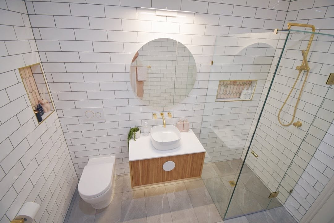 Modern bathroom with white subway tile walls, wooden vanity with round mirror, toilet, and glass shower enclosure