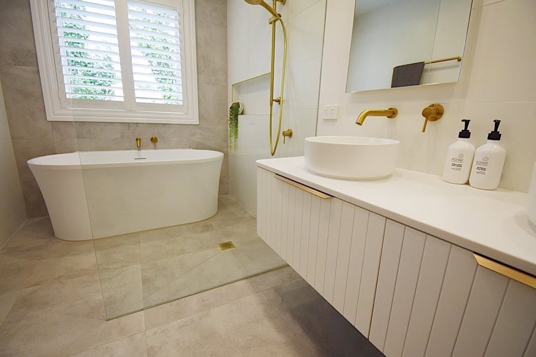 Modern luxury bathroom with freestanding oval bathtub, white vanity with gold fixtures, and stone flooring.