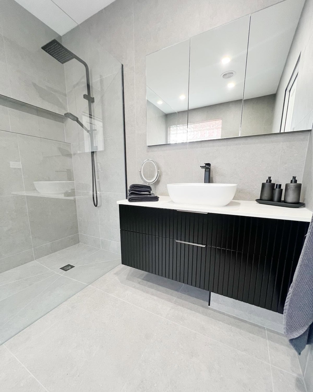 Modern bathroom with black vanity, white vessel sink, large mirror, walk-in shower with glass enclosure, and light tile flooring.