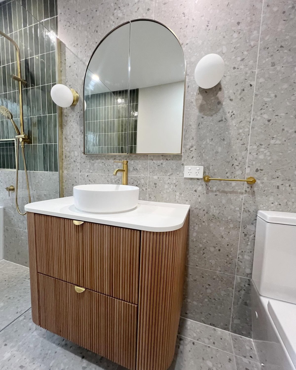 Modern bathroom with round mirror, white vessel sink on wood vanity, terrazzo walls, brass fixtures, and white pendant lights