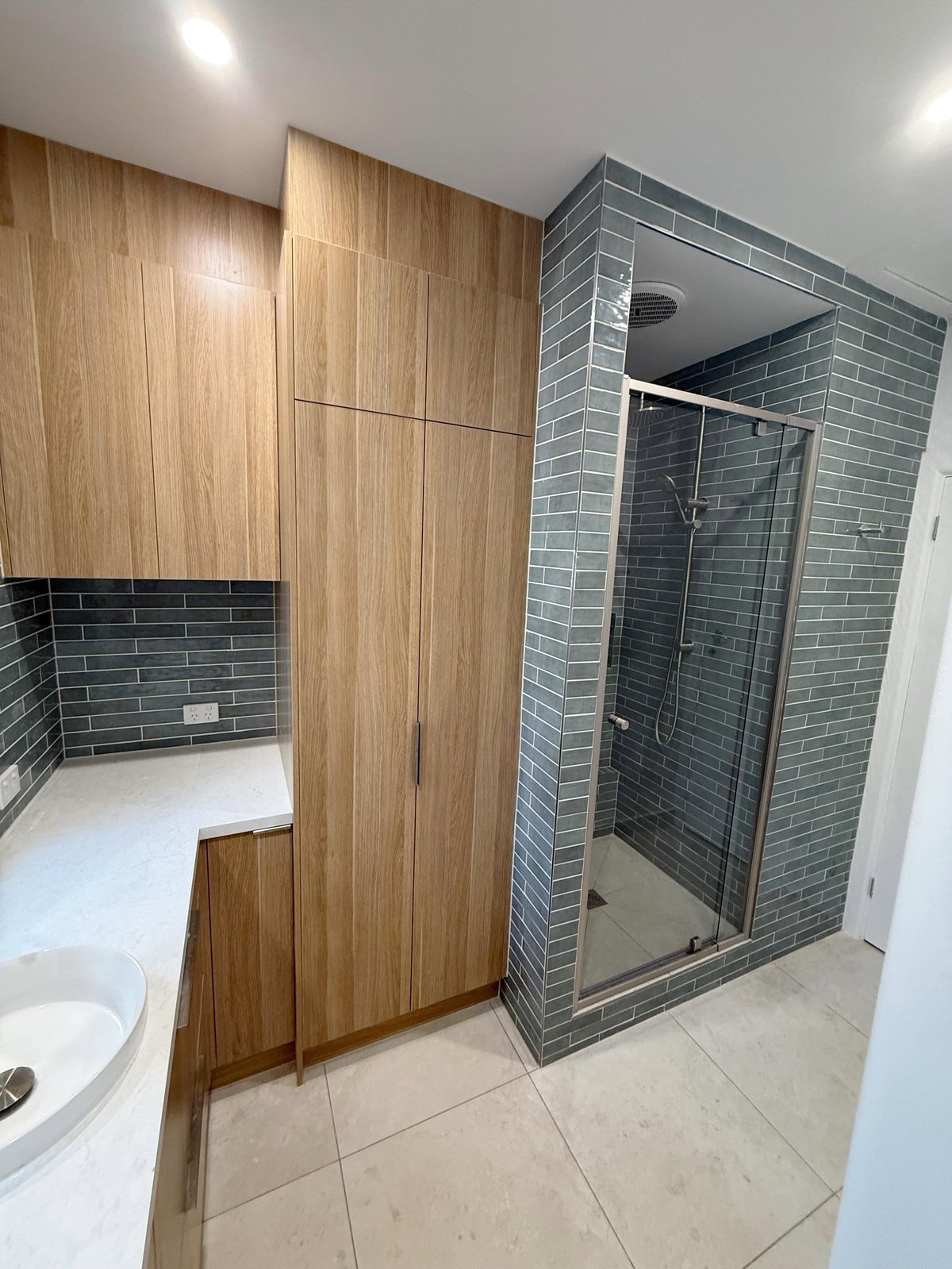 Modern bathroom with wood-paneled wall divider, gray tile shower enclosure, white sink, and beige floor tiles