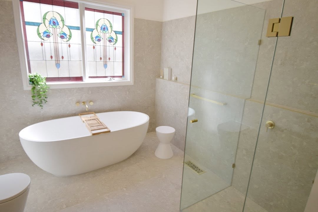 Modern bathroom with white freestanding bathtub, glass shower enclosure, window with decorative panes, and marble flooring