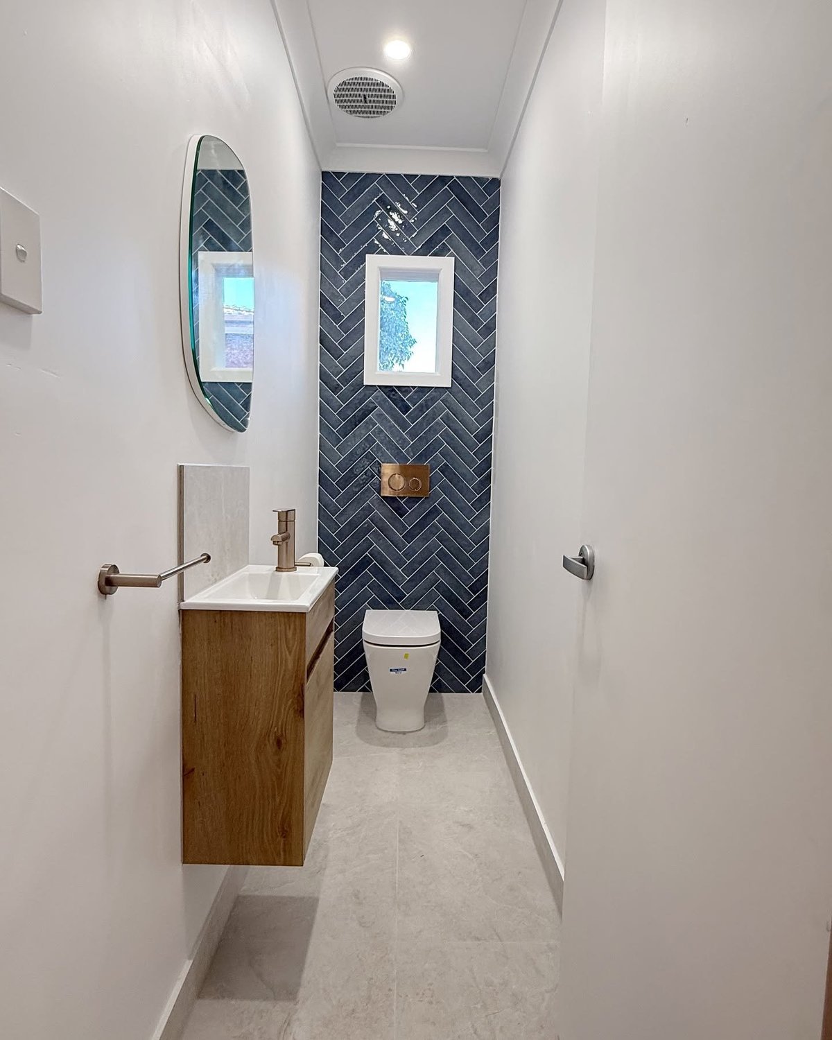 Modern bathroom with wooden vanity, white sink, white toilet, herringbone tile accent wall, and round mirror