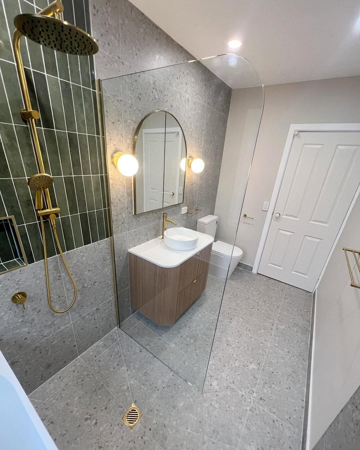Modern bathroom with gray tile walls, glass shower enclosure, floating wood vanity with white sink, arched mirror, and gold fixtures
