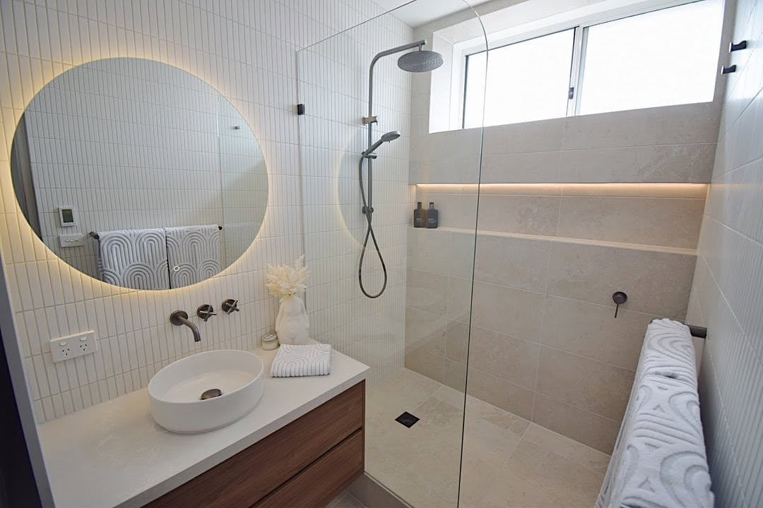 Modern bathroom with round illuminated mirror, white sink, wooden vanity, and glass-enclosed shower with rainfall showerhead and white towel rack