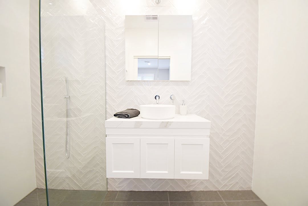 Modern bathroom with white vanity, herringbone tile backsplash, mirror cabinet, and dark floor with glass shower enclosure