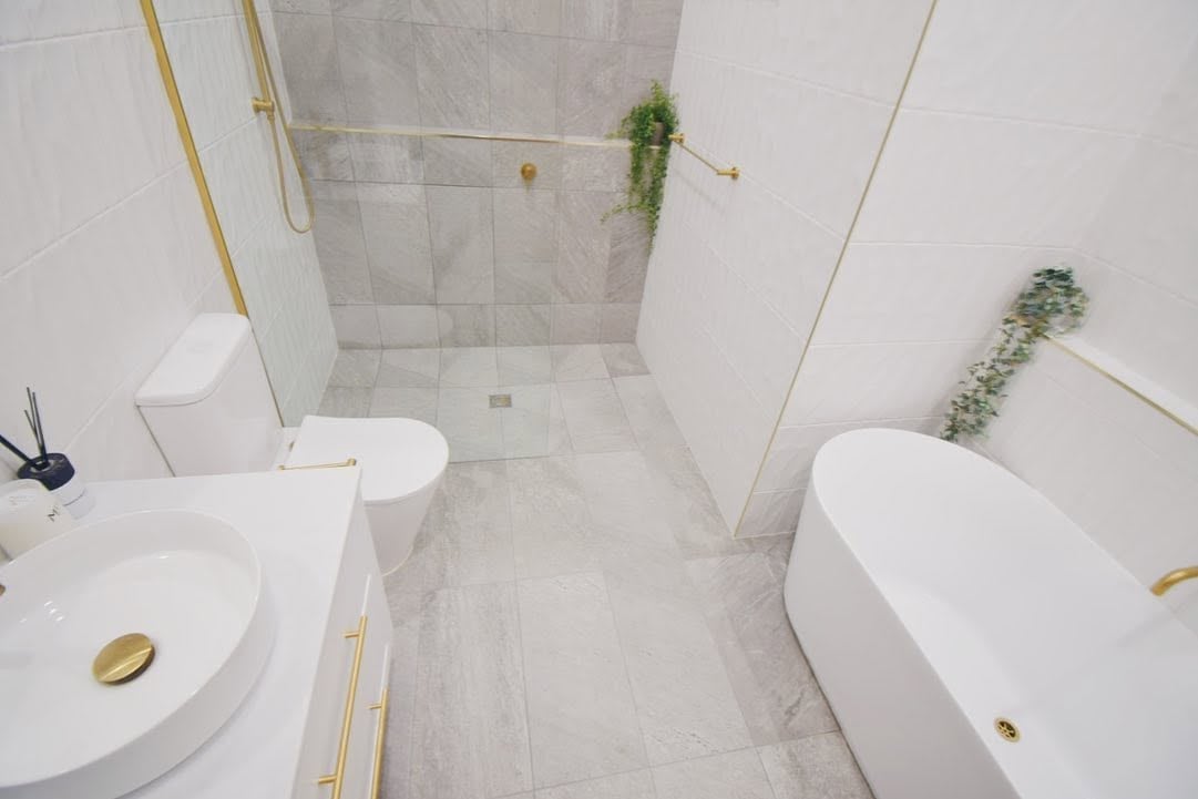 Modern minimalist bathroom with white circular sinks, marble tile flooring, a freestanding bathtub, gold fixtures, and trailing green plants in natural light.