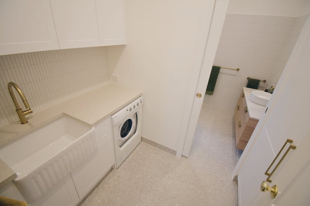 Modern laundry room with white front-loading washer, countertop sink, built-in shelving, and doorway to bathroom